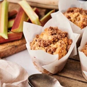 Air Fryer Rhubarb Muffins with crumb topping in paper liners rest on a wooden board, surrounded by fresh rhubarb stalks and two spoons.
