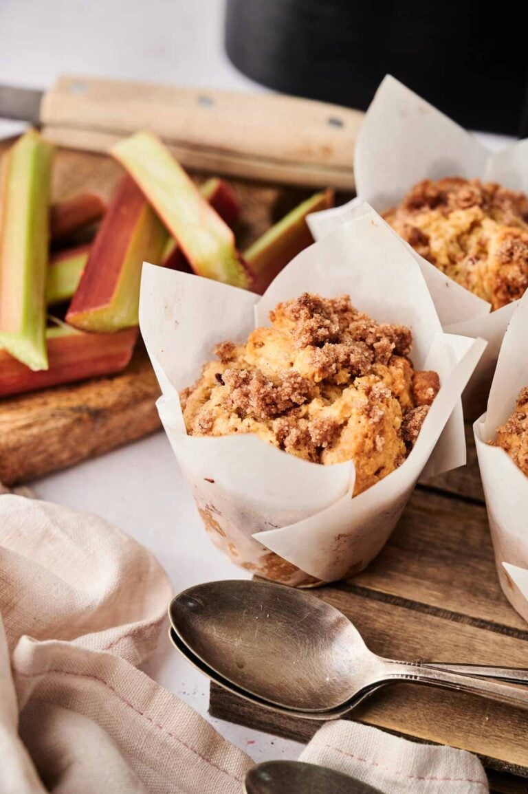 Air Fryer Rhubarb Muffins with crumb topping in paper liners rest on a wooden board, surrounded by fresh rhubarb stalks and two spoons.