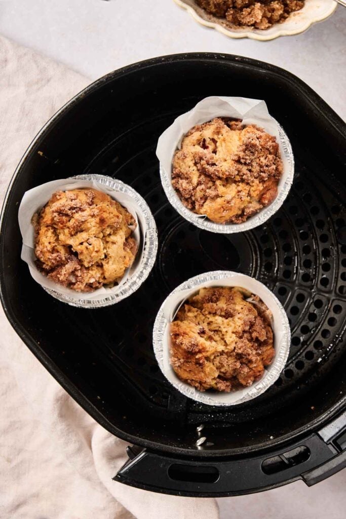 Three crumb-topped Air Fryer Rhubarb Muffins in foil cups rest inside an air fryer basket, perfectly baked and ready to enjoy.