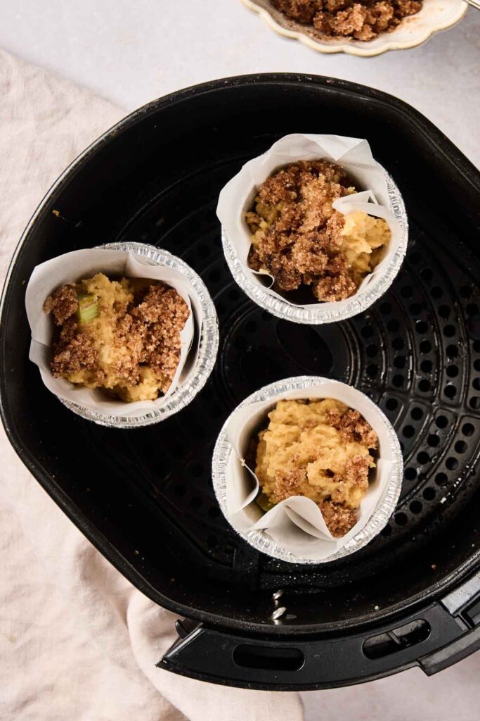 Three unbaked Air Fryer Rhubarb Muffins in cupcake liners with crumb topping are placed inside an air fryer basket, ready to be cooked.