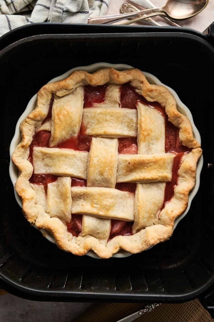 An Air Fryer Rhubarb Pie with a golden lattice crust sits in a white dish on a black tray, accompanied by a cloth and utensils nearby.