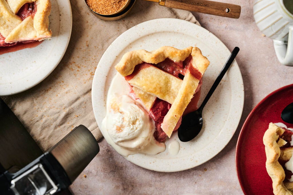 A slice of Air Fryer Rhubarb Pie with lattice crust and a scoop of vanilla ice cream on a plate, accompanied by a black spoon.