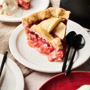 A slice of Air Fryer Rhubarb Pie with a golden lattice crust sits on a white plate next to two black spoons, with another dessert and ice cream visible in the background.