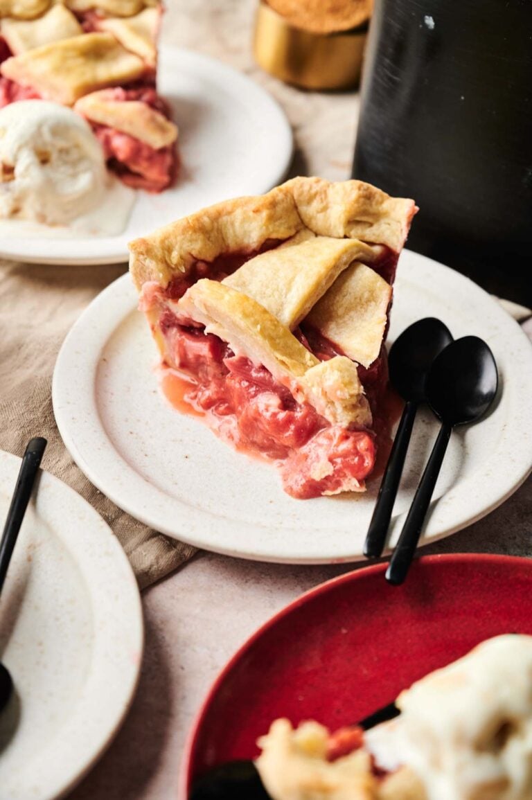 A slice of Air Fryer Rhubarb Pie with a golden lattice crust sits on a white plate next to two black spoons, with another dessert and ice cream visible in the background.