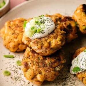Three golden-brown Air Fryer Salmon Patties topped with sour cream, chopped green onions, and dill, served on a beige plate with a fresh basil leaf.