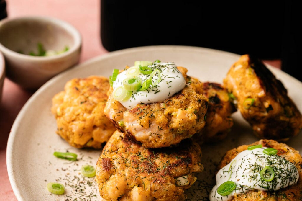 A plate of golden-brown Air Fryer Salmon Patties garnished with a dollop of sour cream, fresh dill, and sliced green onions, with a small bowl in the background.