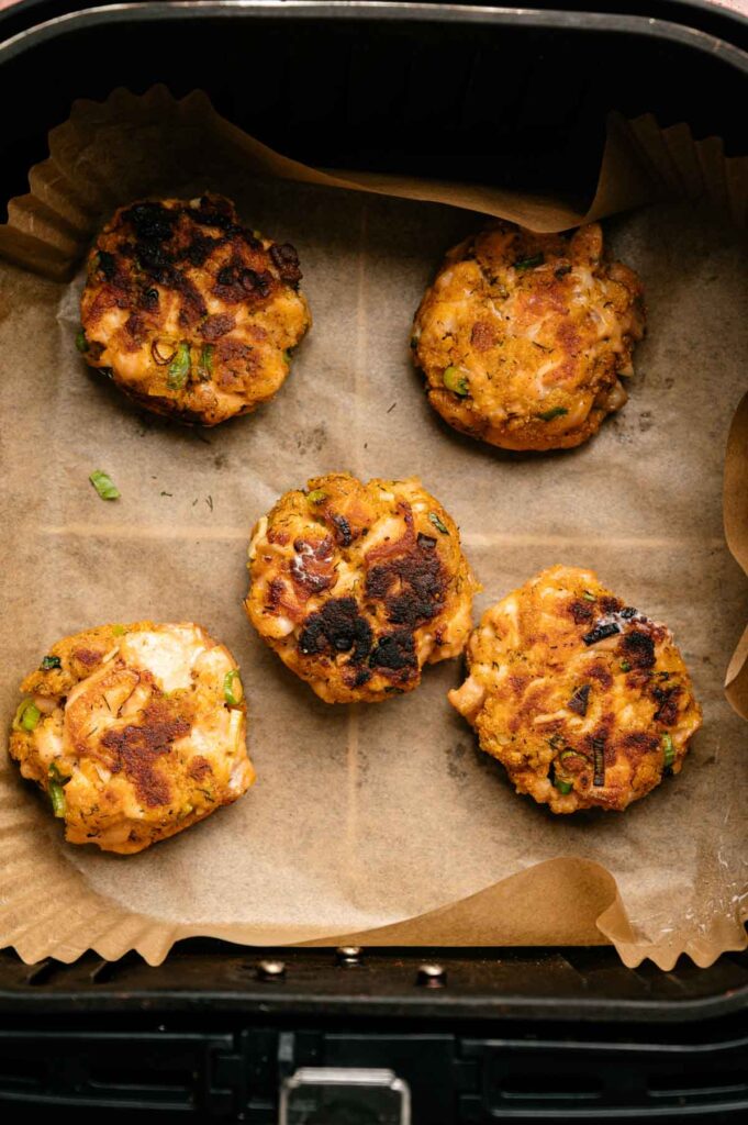 Five browned Air Fryer Salmon Patties are arranged on parchment paper inside an air fryer basket.
