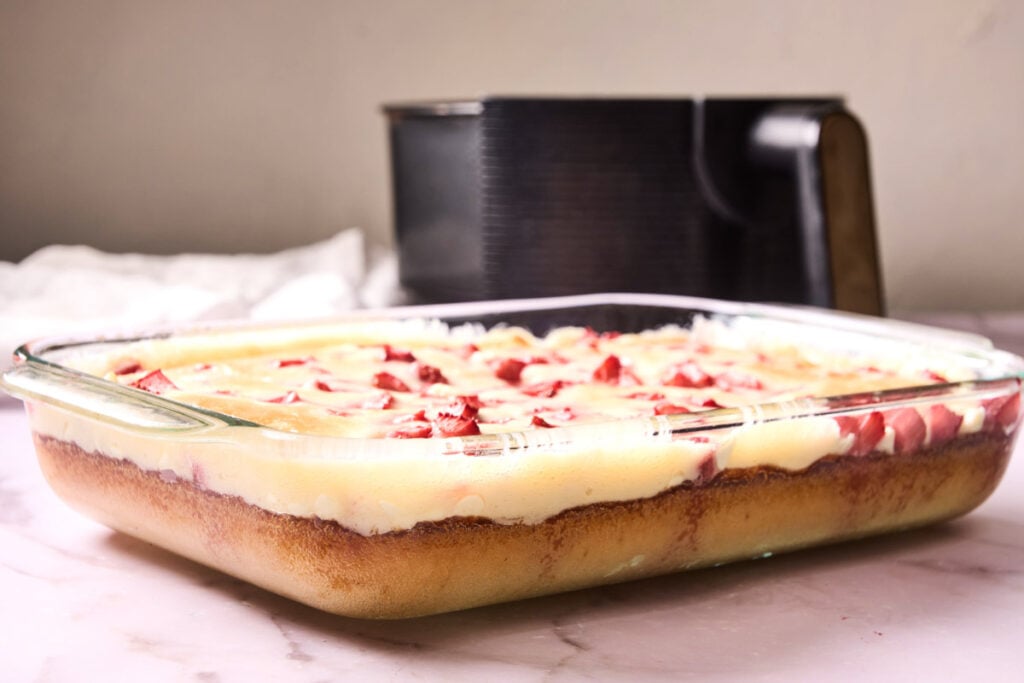 A glass baking dish filled with a layered dessert featuring a golden base, creamy middle layer, and pieces of strawberries on top sits on a marble surface—an inviting presentation reminiscent of a homemade Air Fryer Strawberry Earthquake Cake.
