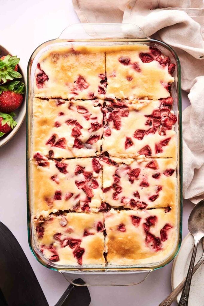 A glass baking dish filled with nine square slices of Air Fryer Strawberry Earthquake Cake bars, each topped with strawberries, is surrounded by a bowl of strawberries and kitchen linens.