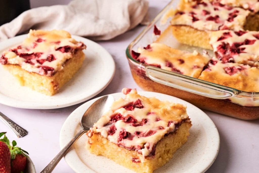 A glass baking dish and plates with slices of yellow Air Fryer Strawberry Earthquake Cake, topped with a white glaze and pieces of strawberries, sit on a light surface.