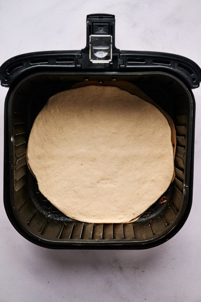 Raw pizza dough placed inside an air fryer basket on a light-colored surface, viewed from above—perfect as the first step to creating your own Air Fryer Taco Pizza.