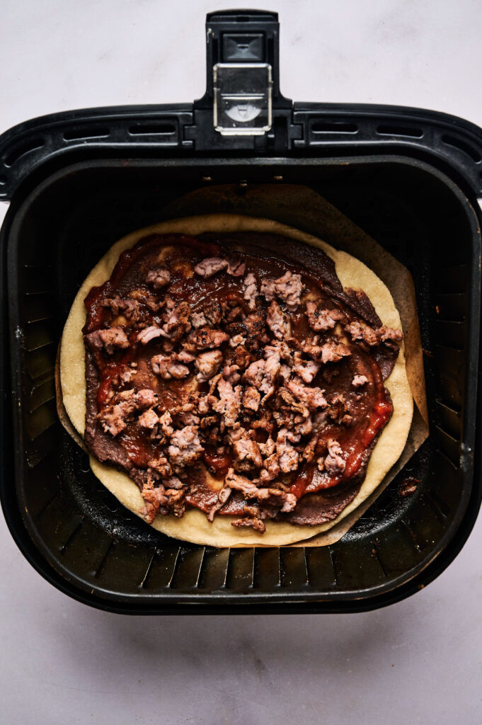 Uncooked Air Fryer Taco Pizza with tomato sauce and crumbled sausage sits in an air fryer basket, ready to be cooked.