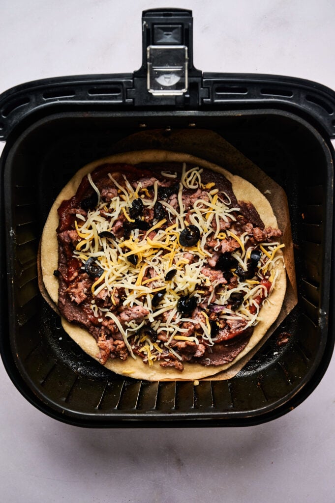 Air Fryer Taco Pizza with ground meat, shredded cheese, and black olives is placed uncooked inside an air fryer basket, ready to be cooked.