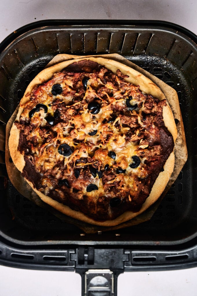 An Air Fryer Taco Pizza with cheese, olives, and toppings sits inside an air fryer basket, ready to be served.