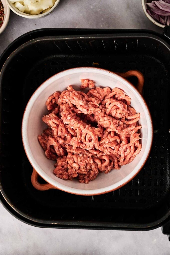 A white bowl filled with raw ground meat sits inside an air fryer basket, ready to be transformed into a delicious Air Fryer Taco Soup, surrounded by small bowls of chopped onions and spices.