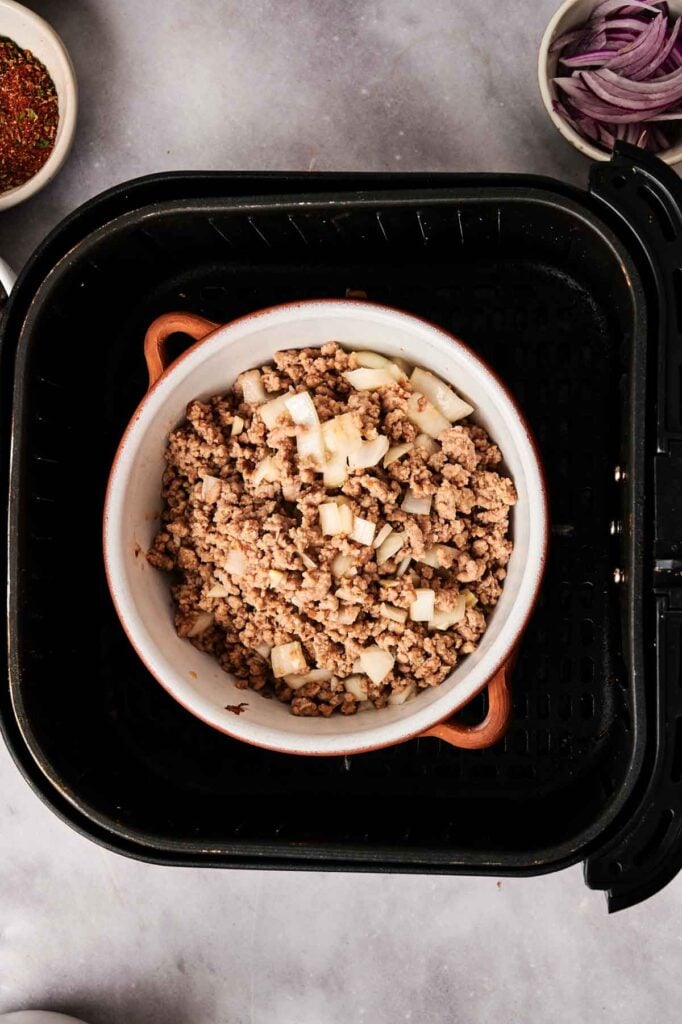 A bowl of cooked ground meat with chopped onions sits inside an air fryer basket, ready to become a flavorful base for Air Fryer Taco Soup, with seasoning and sliced onions in small bowls nearby.