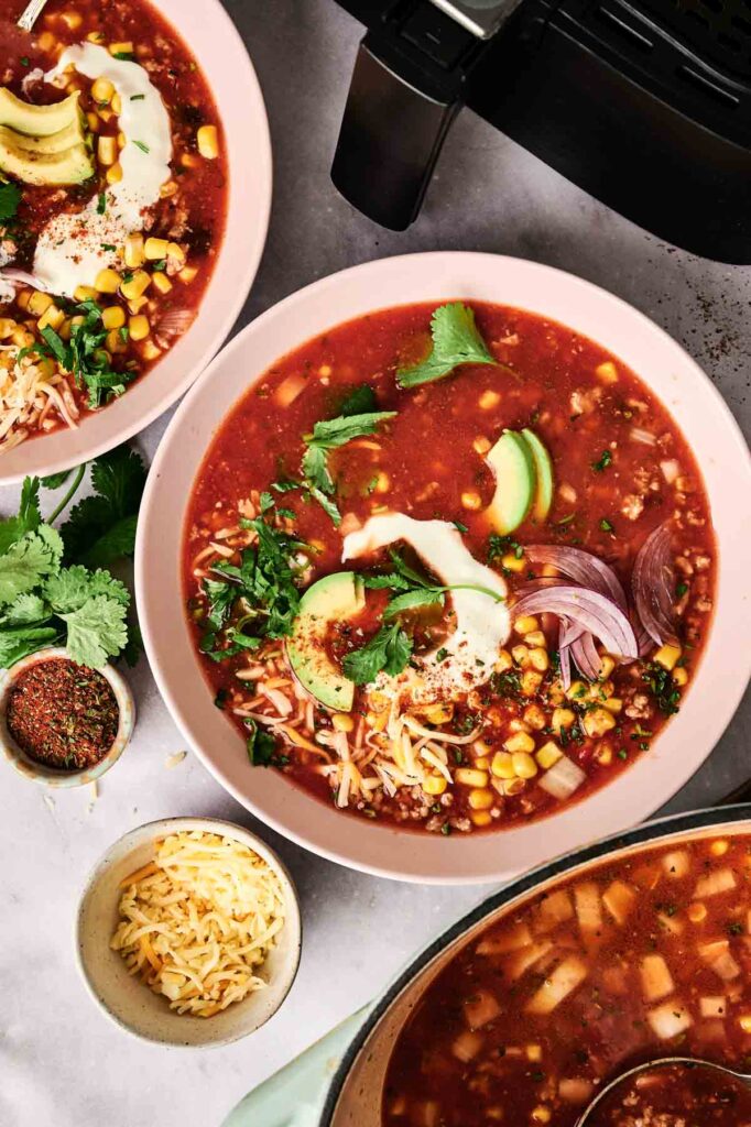Two bowls of Air Fryer Taco Soup with corn, avocado, red onion, cilantro, shredded cheese, and sour cream, next to a pot of soup and small bowls of cheese and spices.