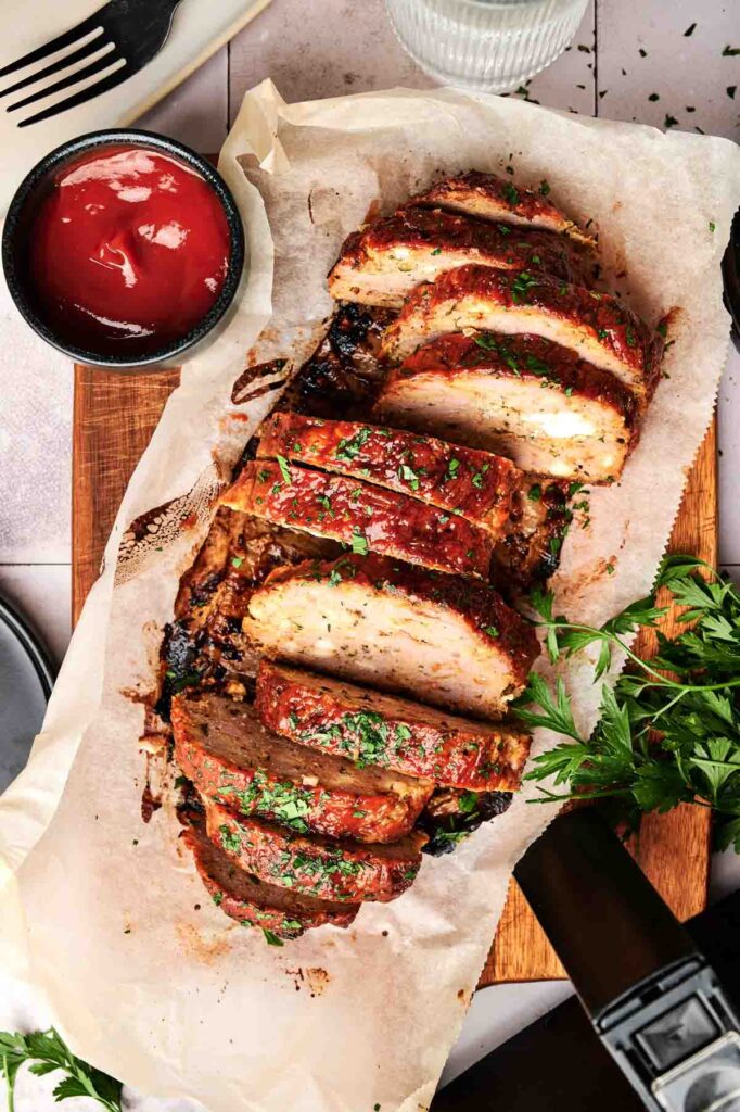 Sliced Air Fryer Turkey Meatloaf garnished with herbs on parchment paper, with a small bowl of ketchup and fresh parsley on the side.