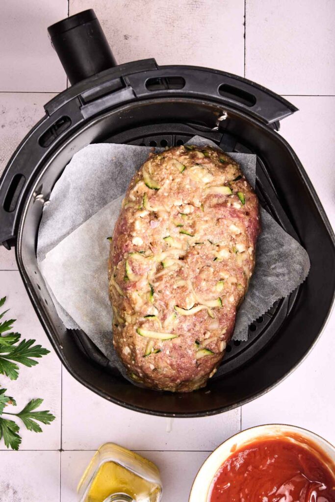 Raw Air Fryer Turkey Meatloaf mixture shaped into a loaf sits on parchment paper inside an air fryer basket, surrounded by small bowls of sauce and herbs on a light tiled surface.