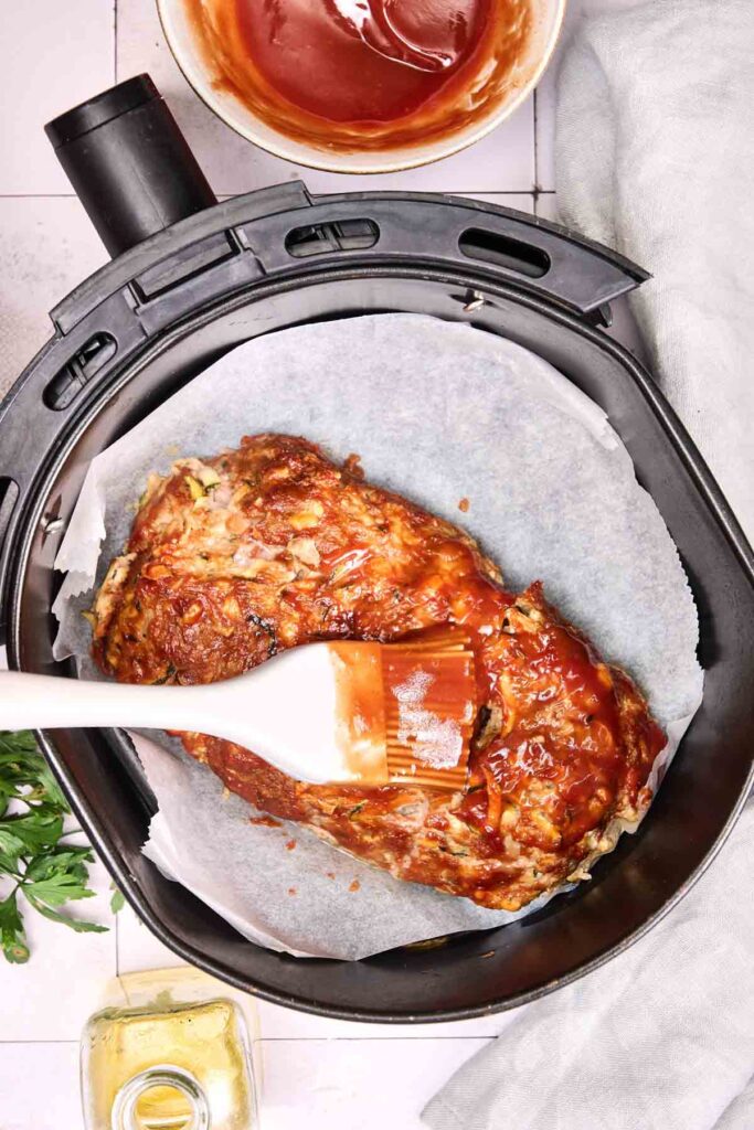 An Air Fryer Turkey Meatloaf sits on parchment paper in an air fryer basket while a hand brushes sauce on top; a bowl of sauce and oil are nearby.