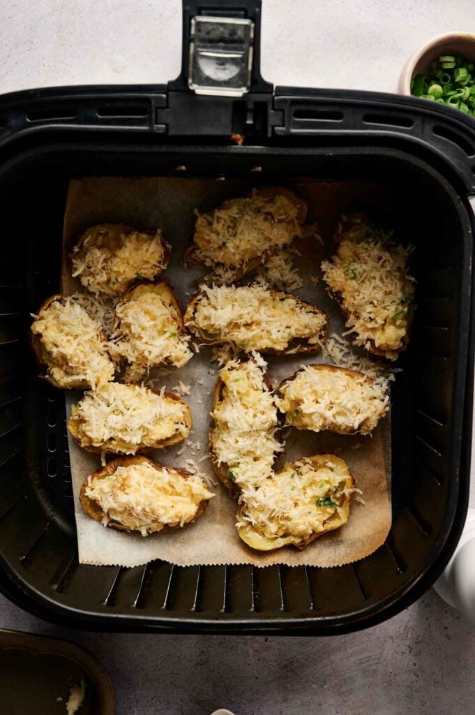 Halved potatoes filled with a creamy cheese mixture and topped with shredded cheese are arranged on parchment paper inside an air fryer basket, ready to become delicious Air Fryer Twice Baked Potatoes.