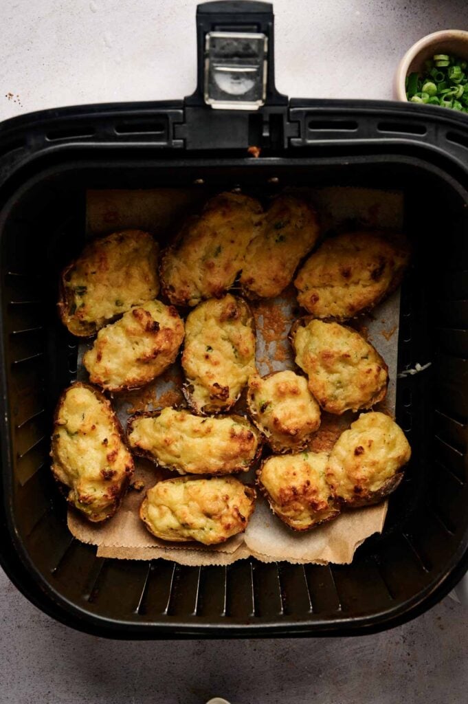 Twelve Air Fryer Twice Baked Potatoes with melted cheese rest on parchment paper inside an air fryer basket. A bowl of chopped green onions is visible in the top right corner.