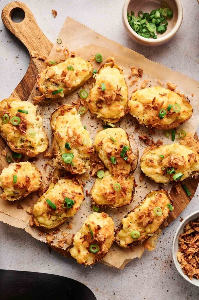 Twelve Air Fryer Twice Baked Potato halves with melted cheese and chopped green onions are arranged on a wooden board lined with parchment paper. Bowls with toppings are nearby.