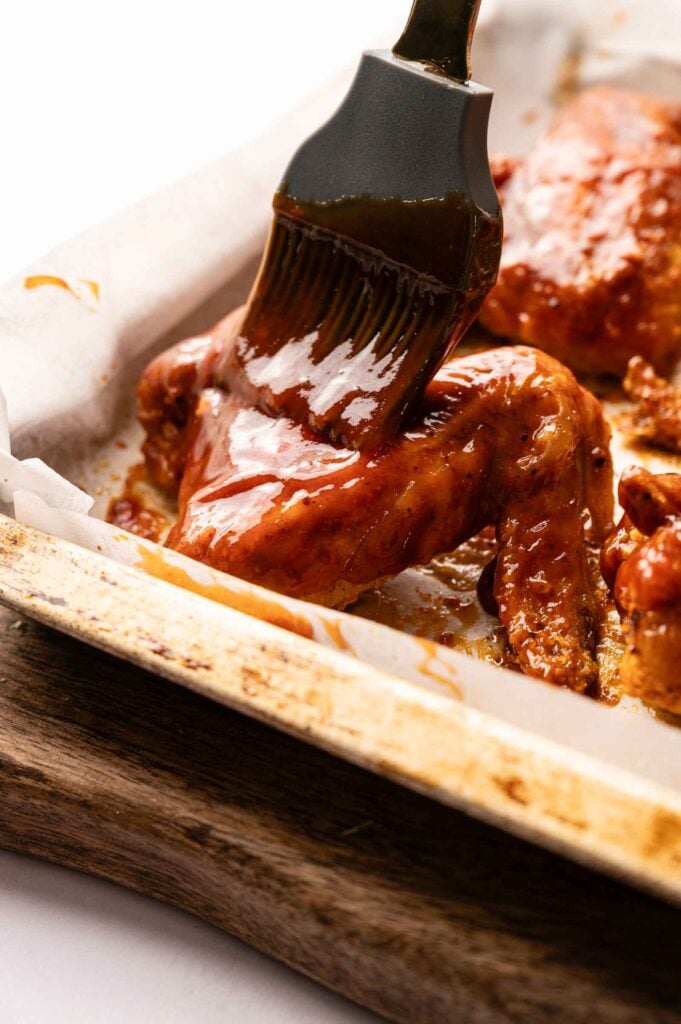 A close-up of a brush applying barbecue sauce to BBQ Wings on a parchment-lined baking tray.