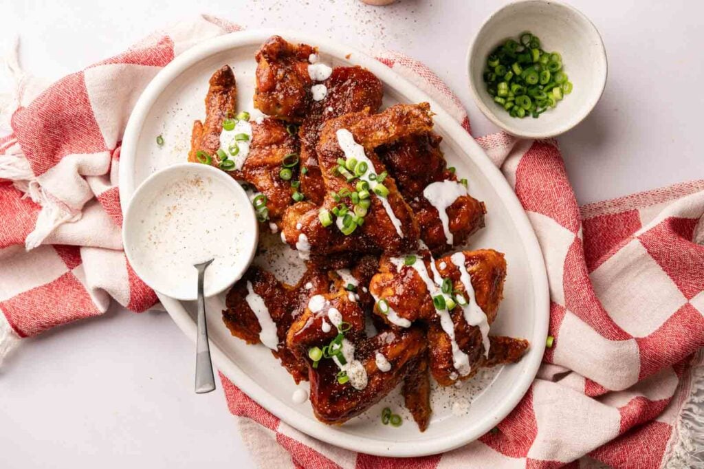 A platter of BBQ wings garnished with chopped green onions and drizzled with white sauce, served alongside a bowl of dip and extra green onions on a red and white cloth.