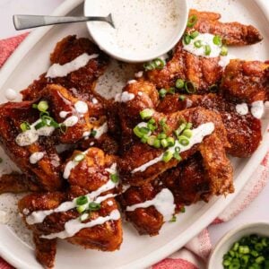 A plate of BBQ Wings garnished with sliced green onions, served with a bowl of creamy dipping sauce on a white platter and a red-and-white cloth.
