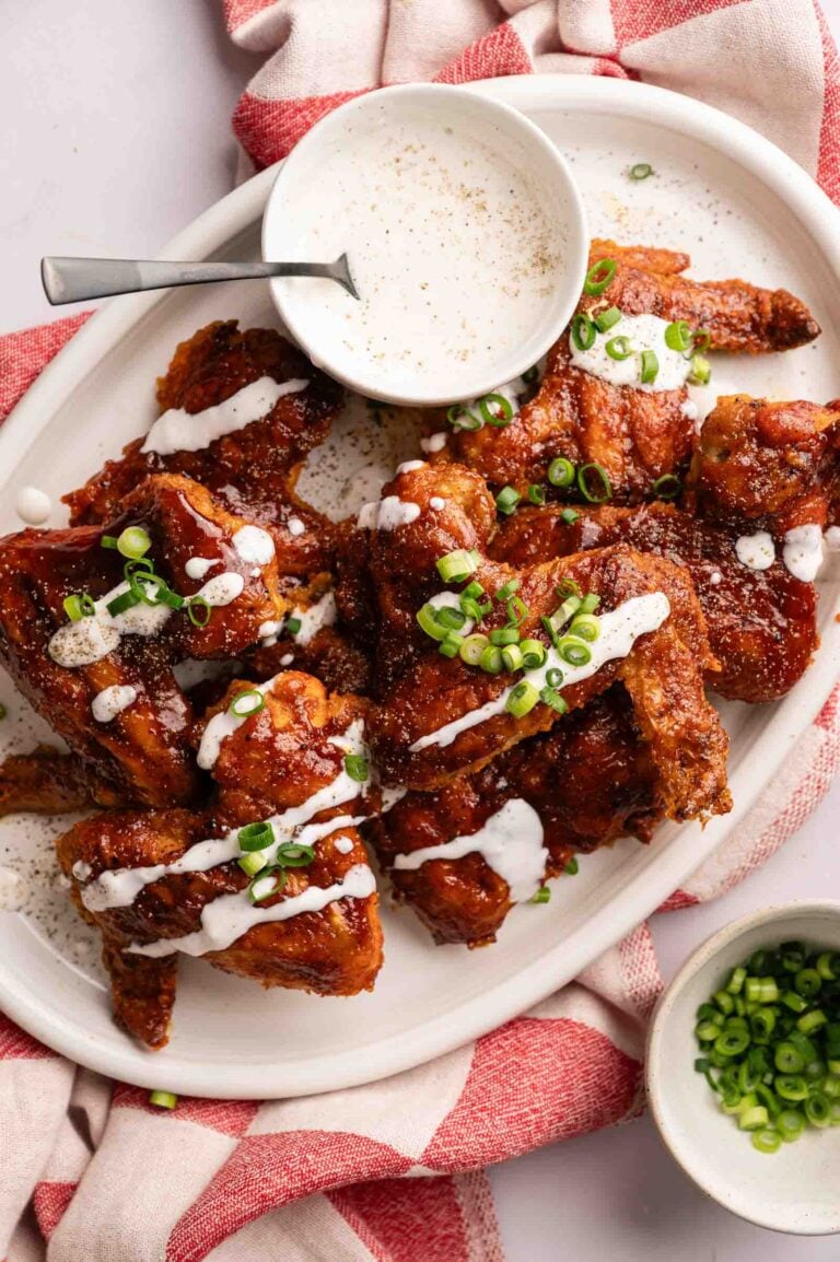 A plate of BBQ Wings garnished with sliced green onions, served with a bowl of creamy dipping sauce on a white platter and a red-and-white cloth.