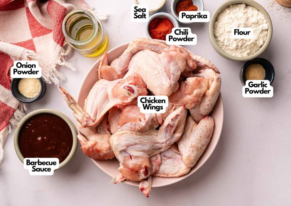A bowl of raw chicken wings, ready to become delicious BBQ Wings, surrounded by small bowls of flour, sea salt, paprika, chili powder, onion powder, garlic powder, barbecue sauce, and a glass of oil.