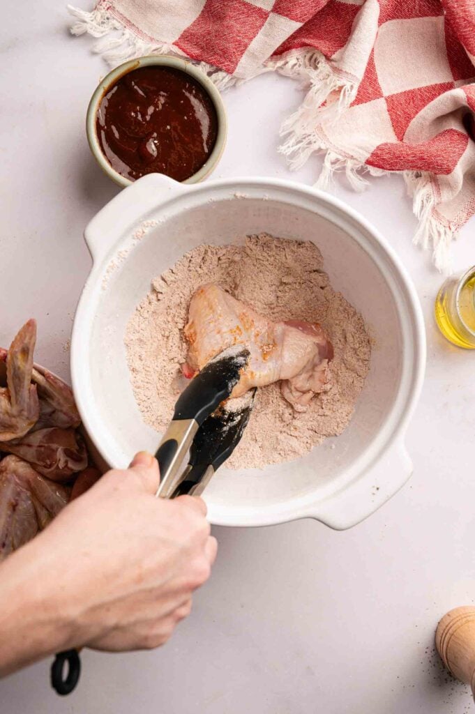 A hand uses tongs to coat a raw chicken wings in seasoned flour in a white mixing bowl, prepping for tasty BBQ wings. Nearby are a bowl of sauce, oil, and a red checked cloth.