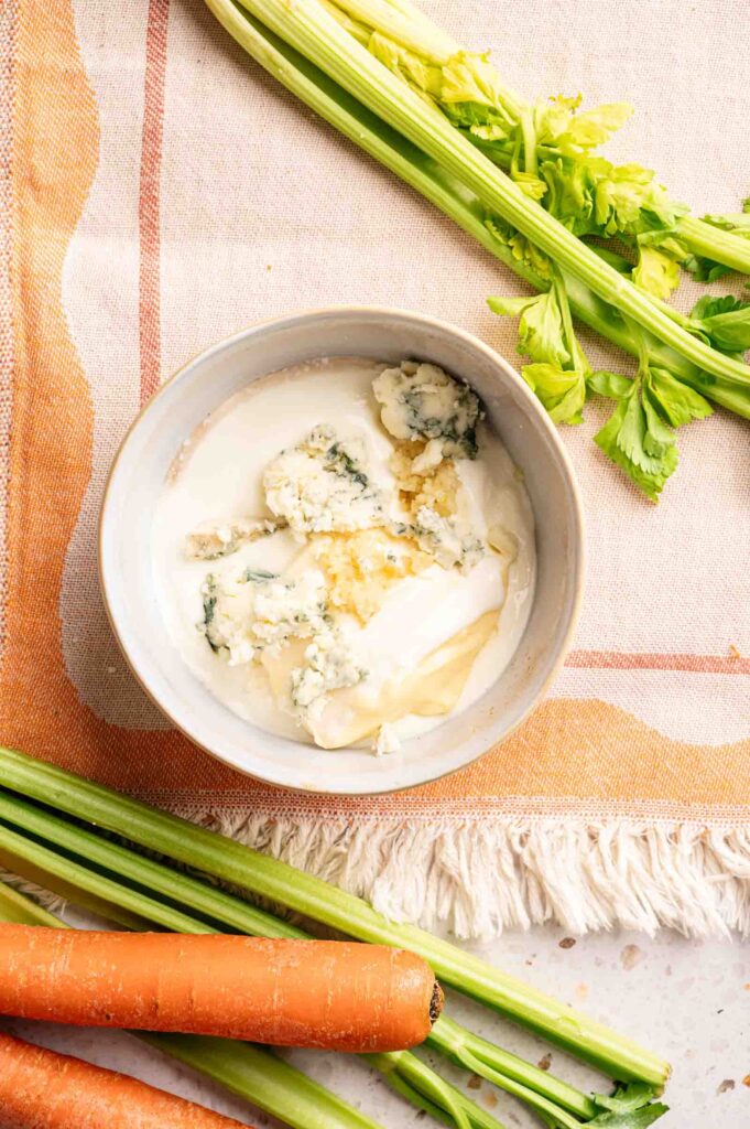 A bowl of blue cheese dressing with crumbled blue cheese and mayonnaise, perfect for dipping Buffalo Wings, surrounded by fresh celery stalks and carrots on a patterned cloth.
