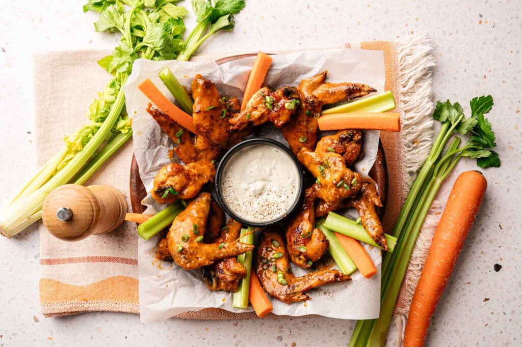 A plate of Buffalo Wings with celery and carrot sticks surrounds a bowl of ranch dip, with fresh celery and a carrot on the table.