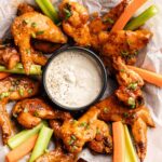 A plate of Buffalo Wings arranged around a bowl of ranch dip, served with celery and carrot sticks on parchment paper.