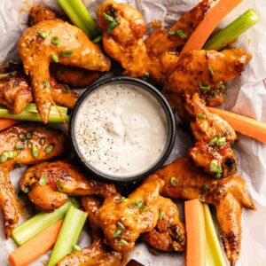 A plate of Buffalo Wings arranged around a bowl of ranch dip, served with celery and carrot sticks on parchment paper.