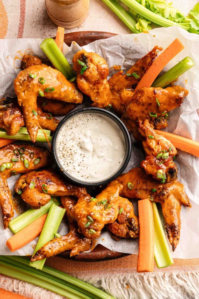 A plate of Buffalo Wings arranged around a bowl of ranch dip, served with celery and carrot sticks on parchment paper.