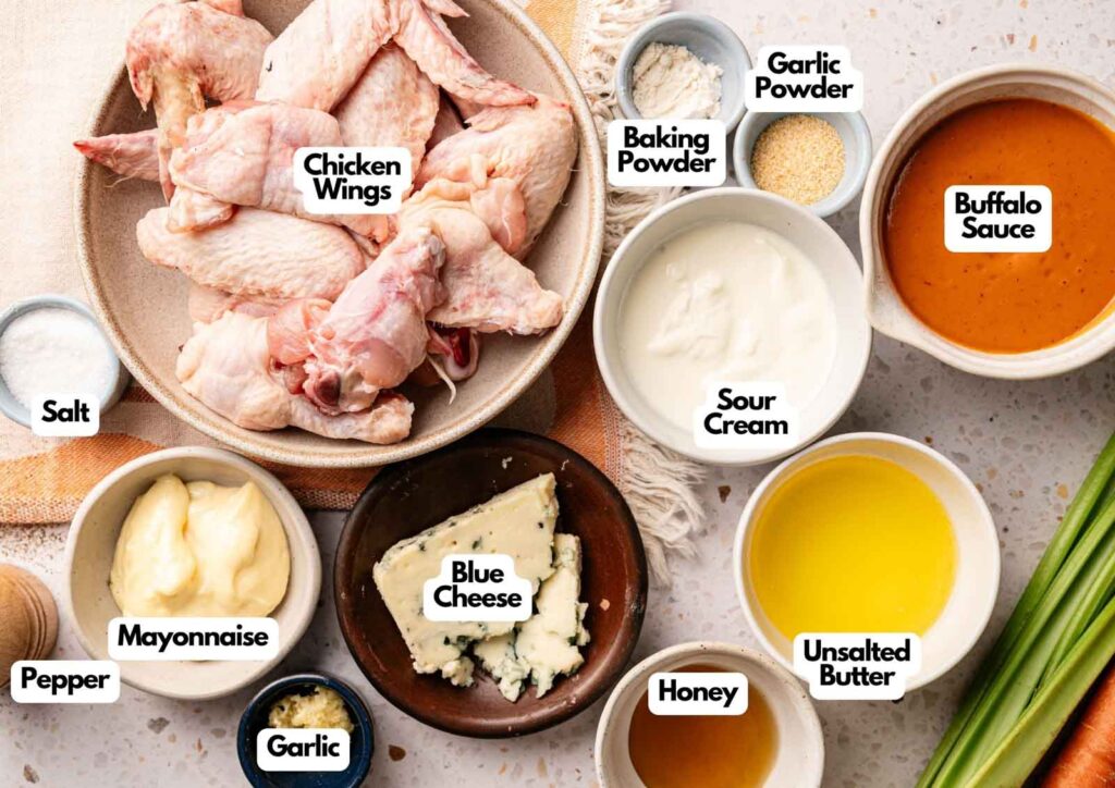 A variety of ingredients for Buffalo Wings are displayed in bowls, including raw chicken wings, sauces, spices, butter, blue cheese, sour cream, and seasonings, each labeled.