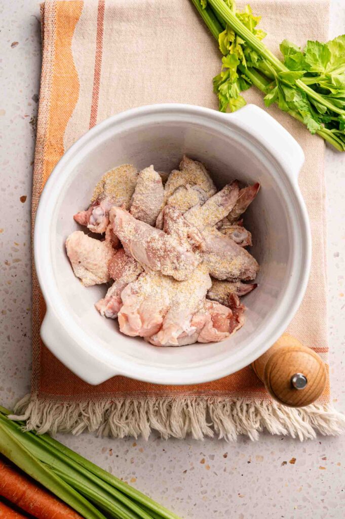 Raw chicken wings coated with seasoning powder in a white bowl, ready to be transformed into classic Buffalo Wings, are placed on a cloth napkin with fresh celery and a pepper grinder nearby.
