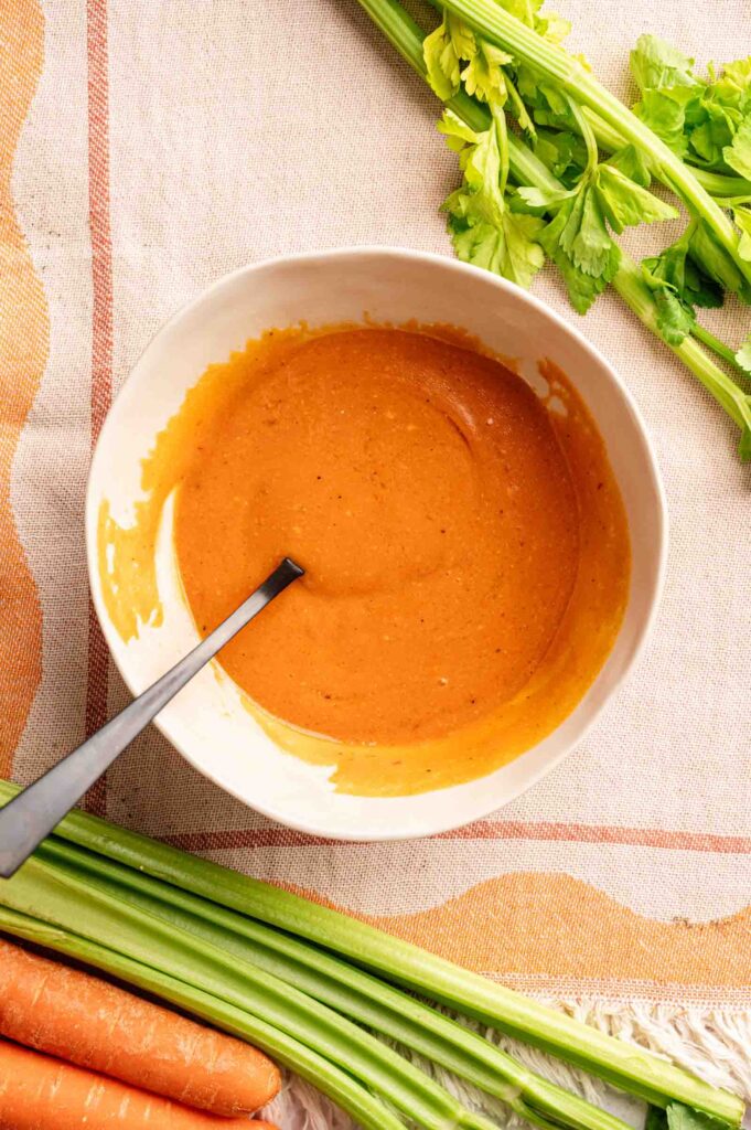 A white bowl of creamy orange buffalo-style dipping sauce with a spoon, surrounded by fresh celery and carrot sticks on a cloth surface.