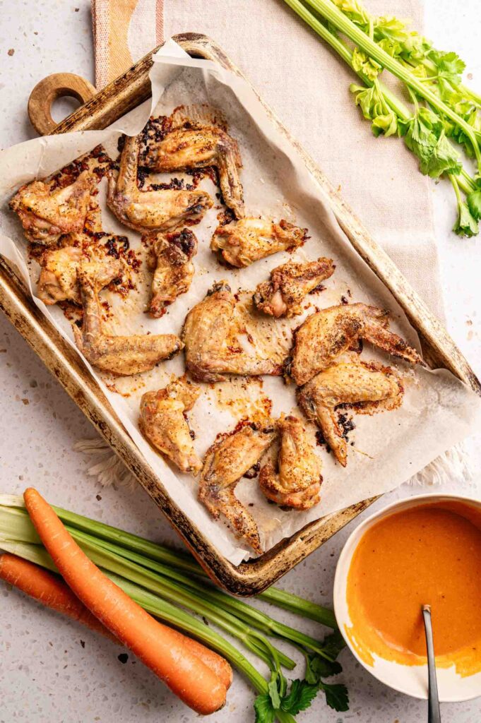 A baking tray with cooked Buffalo Wings on parchment paper, surrounded by celery stalks, carrots, and a bowl of orange dipping sauce with a spoon.