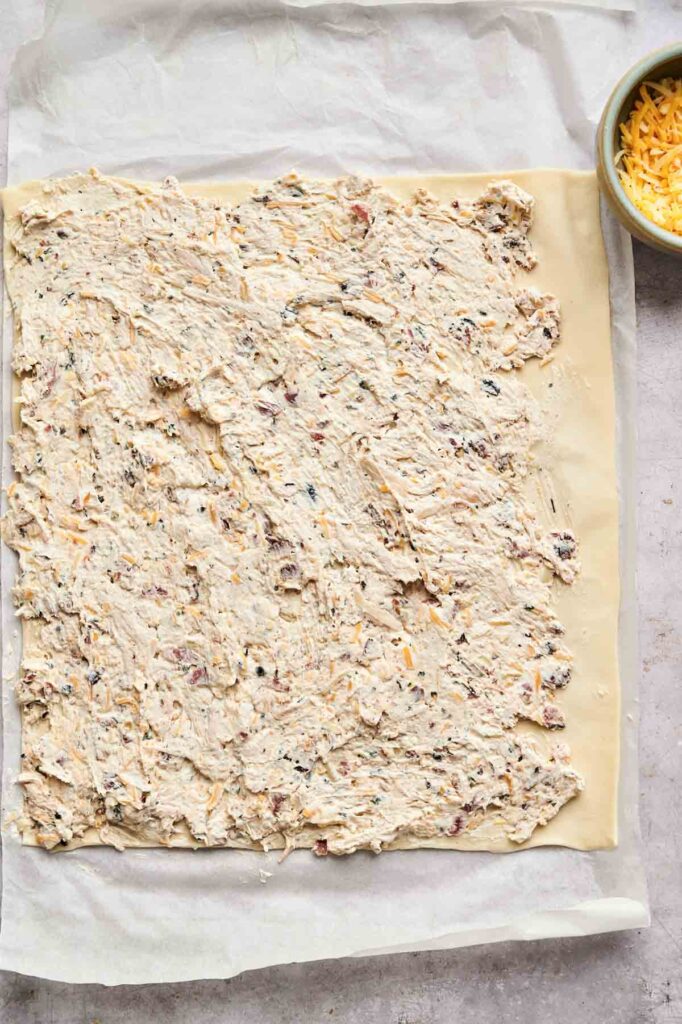 A rectangular sheet of dough on parchment paper is evenly spread with a creamy mixture for Air Fryer Chicken Pinwheels; a small bowl of shredded cheese is visible in the upper right corner.