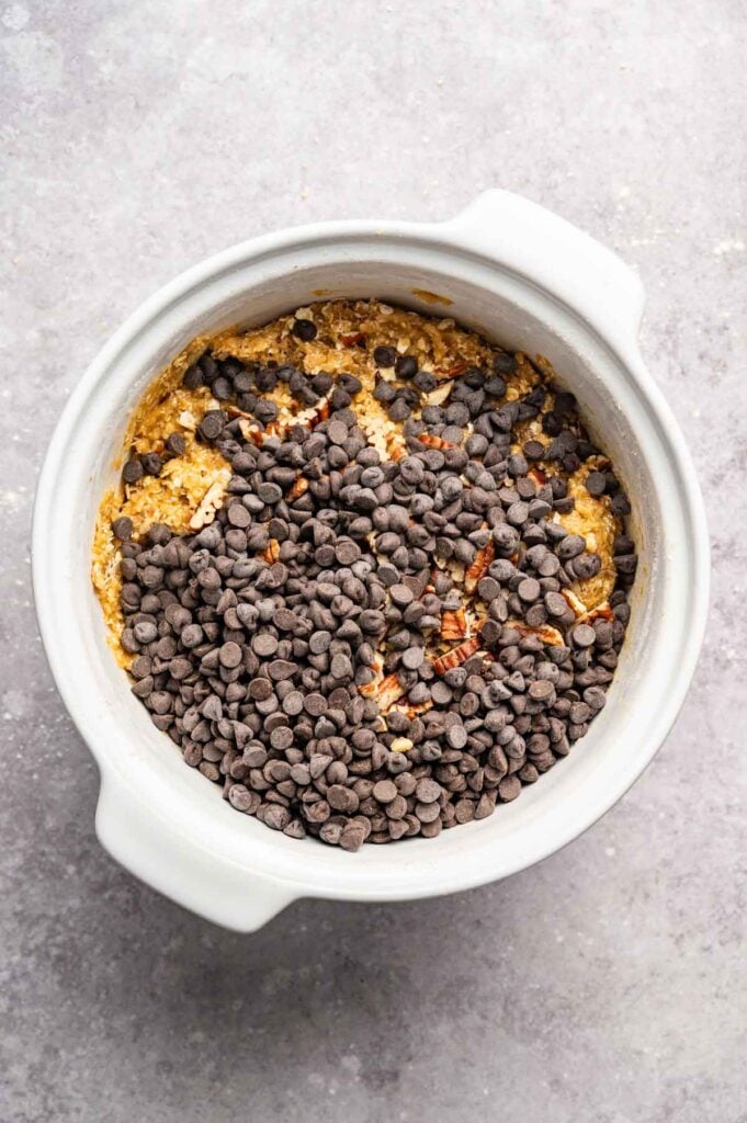 A white mixing bowl filled with Air Fryer Cowboy Cookies dough, chocolate chips, and chopped nuts rests on a gray surface.