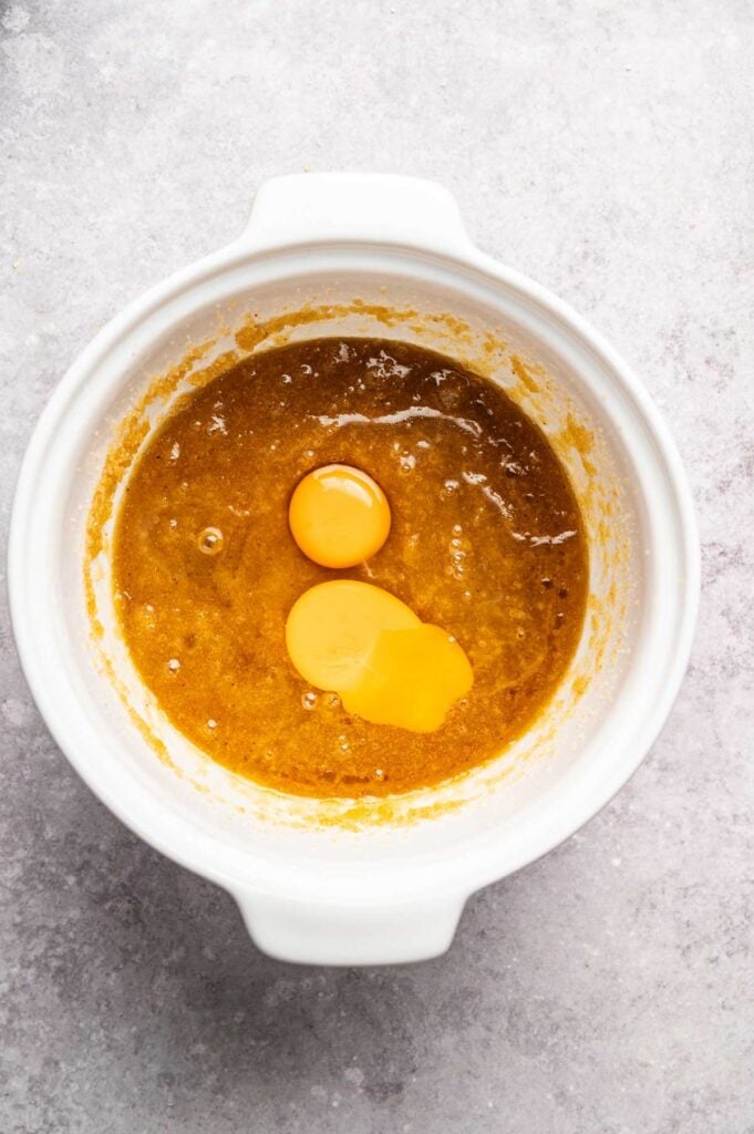 White mixing bowl with a brown batter for Air Fryer Cowboy Cookies and two raw eggs on top, placed on a light gray countertop.