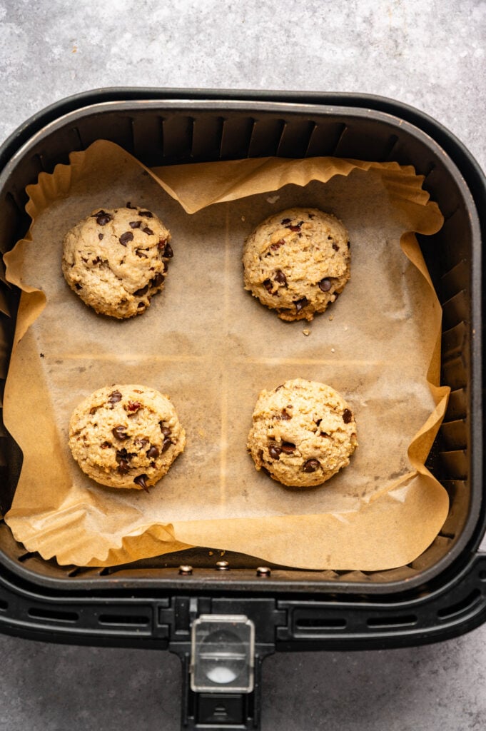 Four scoops of Air Fryer Cowboy Cookies dough, packed with chocolate chips, are placed on parchment paper inside the air fryer basket.