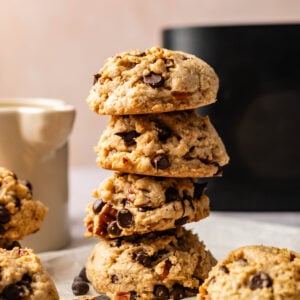 A stack of four Air Fryer Cowboy Cookies sits on a white surface, with loose cookies and chocolate chips nearby; a mug and an air fryer in the background.