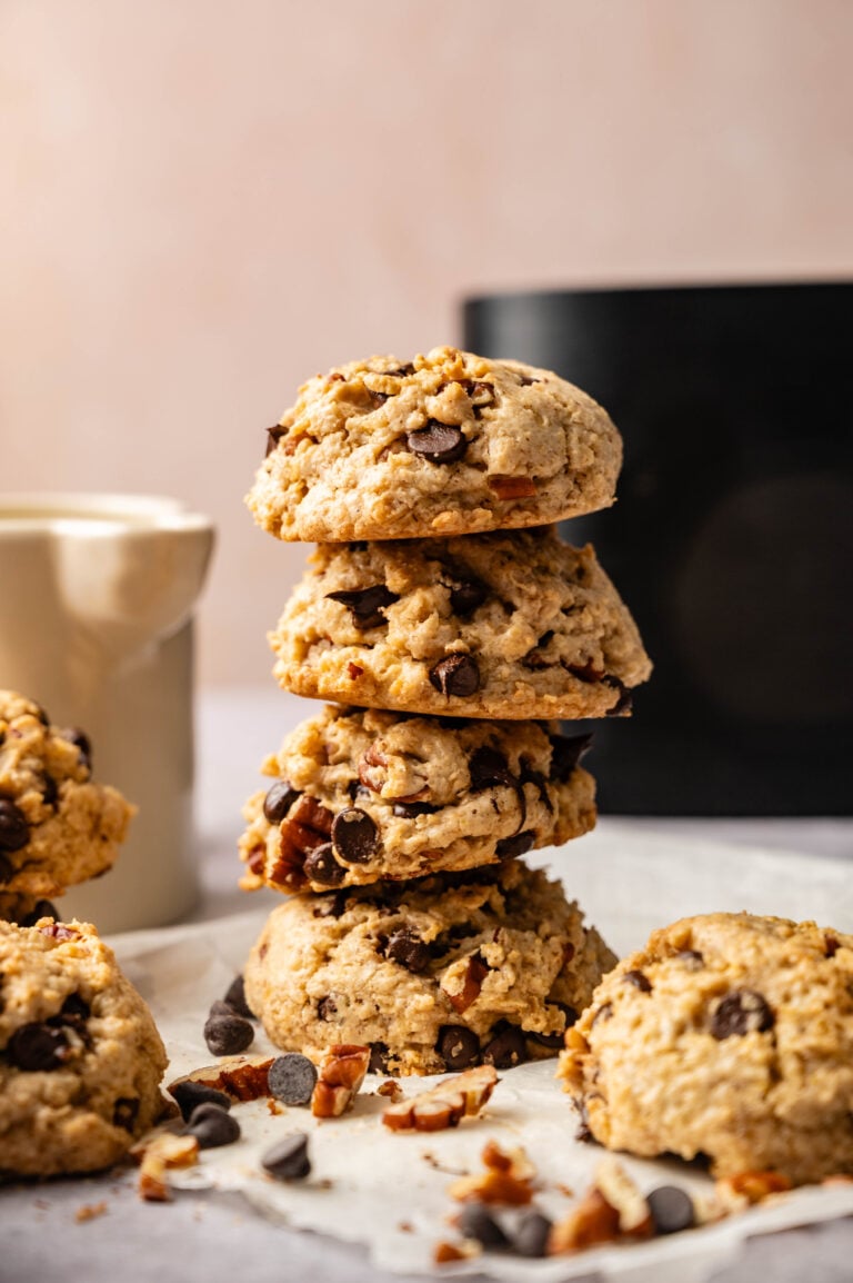 A stack of four Air Fryer Cowboy Cookies sits on a white surface, with loose cookies and chocolate chips nearby; a mug and an air fryer in the background.