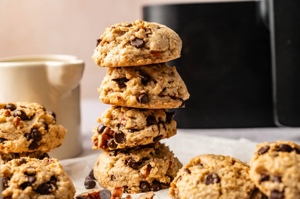 A stack of four Air Fryer Cowboy Cookies sits on a white surface, with loose cookies and chocolate chips nearby; a mug and an air fryer in the background.