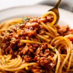 Close-up of a fork twirling spaghetti topped with rich Lamb Bolognese sauce on a white plate.